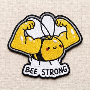 May include: An embroidered patch featuring a yellow and black bee with a smiling face and muscular arms flexed upward. The bee has white wings and is positioned above a white banner with the black text BEE STRONG, all outlined in black thread.