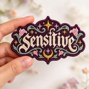 May include: An embroidered patch with a deep purple background featuring the word Sensitive in cream-colored gothic lettering. The design includes light green scrolling vines, pink floral accents, and small yellow and purple stars and a crescent moon.