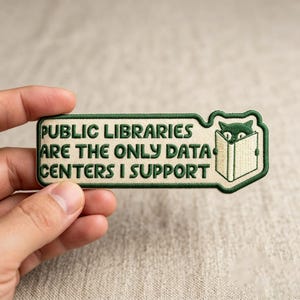 May include: A rectangular embroidered patch with a cream-colored background and dark green border. It features the text "PUBLIC LIBRARIES ARE THE ONLY DATA CENTERS I SUPPORT" in dark green, alongside an illustration of a cat peeking over a book.