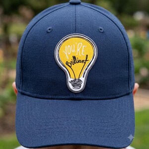 May include: A navy blue baseball cap featuring a central patch shaped like a yellow lightbulb. The patch includes the white text "YOU'RE" and the black cursive text "brilliant" inside the bulb outline, with a black scribbled filament base.