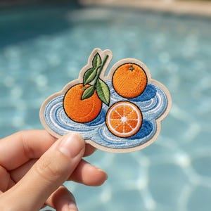May include: An embroidered patch featuring two whole oranges and one orange slice with green leaves, set against a background of blue swirling water ripples. The patch has a light beige border and is shaped to follow the contours of the design.