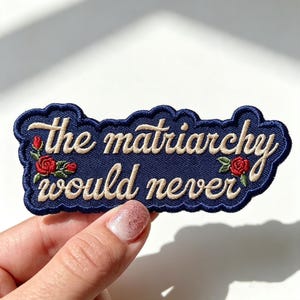 May include: A dark blue embroidered patch with a scalloped edge featuring the text "the matriarchy would never" in cream-colored cursive script, accented by small red roses with green leaves on the left and right sides of the lettering.