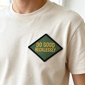May include: A cream-colored short-sleeve t-shirt featuring a diamond-shaped patch on the chest. The patch is olive green with a black border and displays the yellow embroidered text DO GOOD RECKLESSLY in a bold, sans-serif capital font.