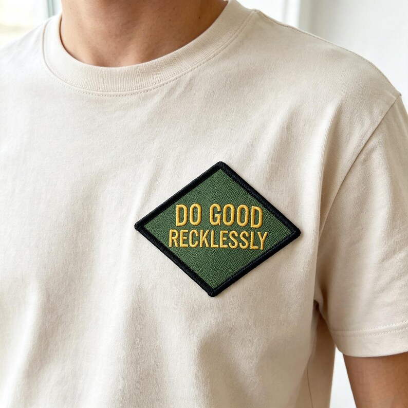 May include: A cream-colored short-sleeve t-shirt featuring a diamond-shaped patch on the chest. The patch is olive green with a black border and displays the yellow embroidered text DO GOOD RECKLESSLY in a bold, sans-serif capital font.