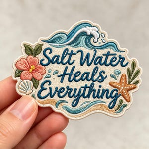 May include: An embroidered patch with a cream background featuring the dark blue text Salt Water Heals Everything. The design includes a teal ocean wave, a pink hibiscus flower, a white seashell, a brown starfish, and green leaves.