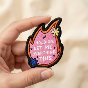 May include: An embroidered patch shaped like a flame with a pink center and orange border. The text "HOLD ON, LET ME OVERTHINK THIS." is stitched in white. It features a small yellow flower at the top, a blue flower at the bottom, and black stars.
