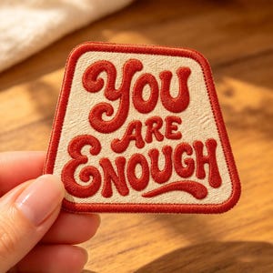 May include: A trapezoidal embroidered patch with a cream-colored background and a thick red border. The patch features the red text "YOU ARE ENOUGH" in a bold, rounded, retro-style font, with the words stacked in three distinct lines.