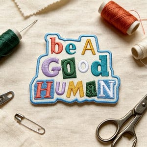 May include: An embroidered patch with a white background and a light blue border featuring the text "be A Good HUMAN" in various colors including red, teal, yellow, purple, green, and blue, surrounded by sewing supplies like thread and scissors.