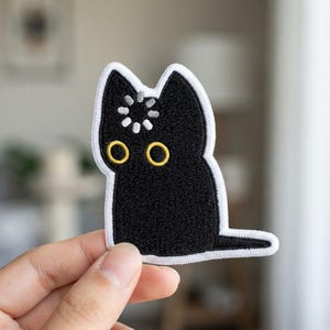 May include: An embroidered patch shaped like a black cat with a white border. The cat has two yellow circular eyes and a white loading icon symbol centered on its forehead. The design features dense black stitching throughout the cat's body.