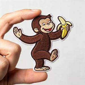 May include: An embroidered iron-on patch featuring a brown monkey with a tan face, smiling while holding a yellow banana. The character has dark brown fur, a red mouth, and is outlined with a thick white border against a plain background.