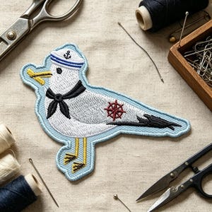 May include: An embroidered patch of a white seagull wearing a sailor hat with an anchor icon, a black neckerchief, and yellow feet. The bird holds a yellow object in its beak and features a red ship's wheel emblem on its side, outlined in light blue.