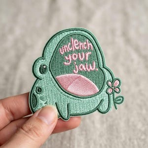 May include: An embroidered patch shaped like a light green frog with a pink mouth and a small pink flower on the side. The patch features the text "unclench your jaw." in pink thread, with a dark green border outlining the entire frog shape.
