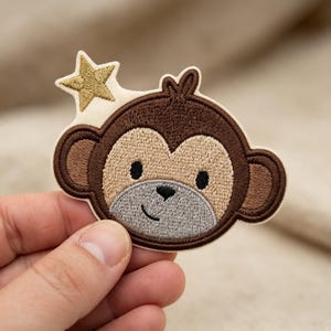 May include: An embroidered patch featuring a brown monkey face with a light tan muzzle, black eyes, and a small black nose. A gold-colored star is positioned above the monkey's head. The patch has a cream-colored border and a stitched texture.