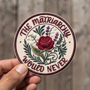 May include: A circular embroidered patch with a cream background and a dark maroon border. The center features a red rose surrounded by green leaves, white lily of the valley, and purple lavender. The text reads THE MATRIARCHY WOULD NEVER.