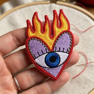 May include: An embroidered patch shaped like a heart with a central blue eye, surrounded by yellow flames with red outlines. The heart is light purple with green stitched details around the edges and a white sclera around the blue iris.