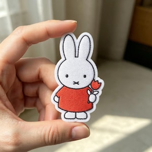 May include: An embroidered iron-on patch featuring a white rabbit character with long ears, black eyes, and a black X-shaped mouth. The rabbit wears a solid red shirt and holds a small red tulip flower with green leaves in its hand.