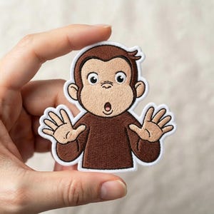 May include: An embroidered iron-on patch featuring a brown monkey with a tan face, large eyes, and an open mouth expression. The monkey has both hands raised with palms facing forward and is outlined with a thick white border for easy application.