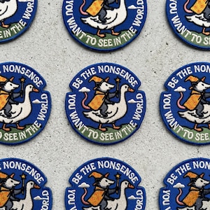 May include: A circular embroidered patch featuring a white goose carrying a grey opossum wearing a yellow wizard hat and cloak. The opossum holds a star-tipped wand. The blue border contains the text BE THE NONSENSE YOU WANT TO SEE IN THE WORLD.