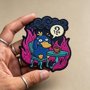 May include: An embroidered patch featuring a blue duck-like creature sitting at a teal table with a brown cup. The scene is surrounded by bright pink flames and a dark blue cloud above. A speech bubble contains a black question mark symbol.