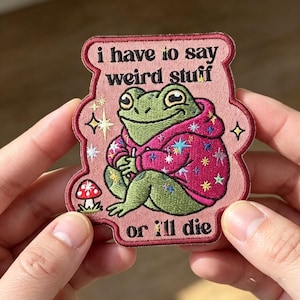 May include: An embroidered patch with a dusty pink background featuring a green frog wearing a bright pink hoodie decorated with colorful stars. The text reads "i have to say weird stuff or i'll die" in black, with a small red and white mushroom.
