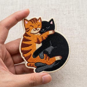 May include: An embroidered patch featuring an orange tabby cat and a solid black cat curled together in a hug. The cats have pink inner ears and are depicted with closed eyes. The patch has a cream-colored border and a textured fabric finish.