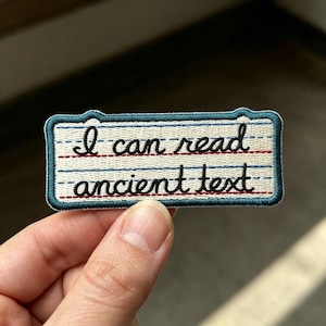 May include: A rectangular embroidered patch featuring a cream-colored background with blue and red horizontal lines resembling notebook paper. The black embroidered text reads "I can read ancient text" in a cursive script with a dark teal border.