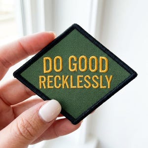 May include: A diamond-shaped embroidered patch with an olive green background and a thick black border. The text DO GOOD RECKLESSLY is stitched in bold yellow capital letters across the center of the patch, creating a clear contrast.