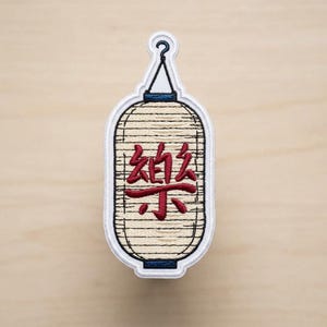 May include: An embroidered patch shaped like a traditional Japanese lantern. The lantern is cream-colored with horizontal black stitching, featuring a dark red kanji character in the center. It has a blue top and bottom trim with a black hook.