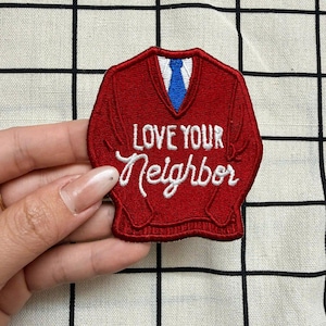 May include: A red embroidered patch shaped like a V-neck sweater with a white shirt collar and a blue necktie visible at the neckline. The text LOVE YOUR Neighbor is stitched in white thread across the center of the red sweater patch.