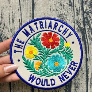 May include: A circular embroidered patch with a cream-colored background and a blue border. The text "THE MATRIARCHY WOULD NEVER" is stitched in blue around the edge. The center features a floral arrangement with red, yellow, and teal flowers.