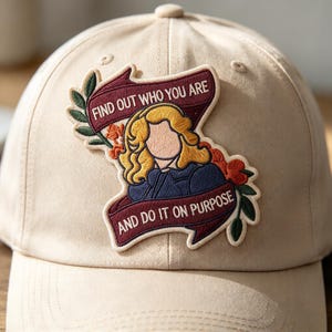 May include: A beige baseball cap featuring an embroidered patch of a blonde figure with no facial features, surrounded by orange flowers and green leaves. Two maroon banners contain the text: FIND OUT WHO YOU ARE AND DO IT ON PURPOSE.