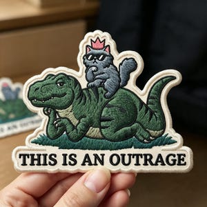 May include: An embroidered patch featuring a green Tyrannosaurus rex lying on grass with a grey cat wearing a pink crown and black sunglasses sitting on its back. The text THIS IS AN OUTRAGE is embroidered in black at the bottom of the patch.