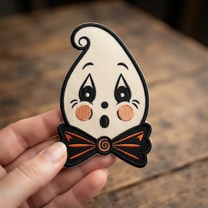 May include: An embroidered patch shaped like a ghost with a cream-colored body, black outlines, and a black bow tie featuring orange accents. The ghost has large black eyes, a small round mouth, and circular peach-colored cheeks on its face.