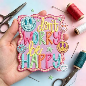 May include: A rectangular fabric patch with a scalloped pink border featuring the embroidered text "don't worry be HAPPY" in yellow, pink, green, and blue. The design includes five small smiley face icons with various patterns and colors.