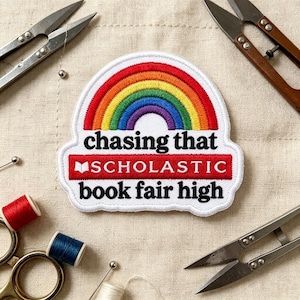May include: A white embroidered patch featuring a colorful rainbow arch above the black text "chasing that" and "book fair high" with the word "SCHOLASTIC" in white letters on a red rectangular background with a small book icon.