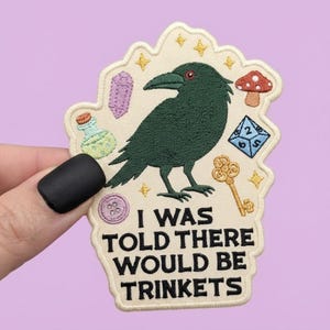 May include: An embroidered patch with a cream background featuring a dark green crow, a purple crystal, a green potion bottle, a red mushroom, a blue die, a gold key, and a purple button. The text reads: I WAS TOLD THERE WOULD BE TRINKETS.