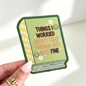 May include: A rectangular embroidered patch shaped like a book with a light green cover, a yellow and white checkered spine, and white page edges. The text reads THINGS I WORRIED ABOUT THAT TURNED OUT JUST FINE, accented by two yellow smiley faces and white flowers.