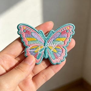 May include: An embroidered butterfly patch featuring a vibrant color palette of pink, teal, yellow, and lavender threads. The design includes a teal body and a white script text overlay that reads Polly, all outlined with a thick teal border.