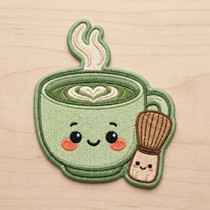 May include: An embroidered patch featuring a light green cup of matcha tea with a smiling face, pink cheeks, and white steam rising from the top. A small, tan bamboo matcha whisk with a smiling face is positioned next to the cup.