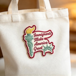 May include: A cream-colored canvas tote bag featuring an embroidered patch. The patch depicts a green Statue of Liberty torch with a yellow flame and a green crown, alongside the red embroidered text: Immigrants Make America Great.