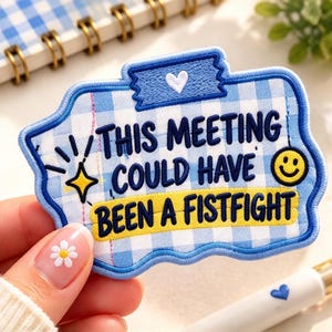 May include: A rectangular embroidered patch with a light blue and white gingham pattern. It features the dark blue text 'THIS MEETING COULD HAVE BEEN A FISTFIGHT' with a yellow smiley face, a yellow star icon, and a small white heart.