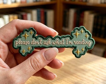 May include: An embroidered patch in a sage green shade with a dark green scalloped border. The patch features gold star accents and the text "please don't ask what I'm reading" stitched in a light cream thread across the center of the design.
