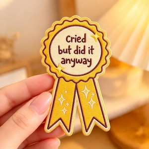 May include: A yellow embroidered award ribbon patch with a scalloped edge and two hanging tails. The center features the dark brown text Cried but did it anyway, accented by small white and yellow embroidered star shapes throughout.