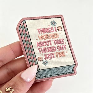May include: An embroidered patch shaped like a book with a cream-colored cover, pink border, and teal spine. The text reads, "THINGS I WORRIED ABOUT THAT TURNED OUT JUST FINE," accented with small embroidered pink smiley faces and light blue flowers.