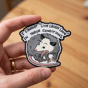 May include: An embroidered patch featuring a gray and white opossum with an open mouth, surrounded by a black border. The text above the opossum reads, "I CANNOT "LIVE LAUGH LOVE" IN THESE CONDITIONS." The patch has a white background and black stitching.