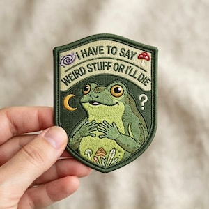 May include: An embroidered patch with a dark green border featuring a light green frog, a crescent moon, a spiral, a question mark, and mushrooms. The text reads: I HAVE TO SAY WEIRD STUFF OR I'LL DIE. The patch has a shield-like shape.