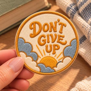 May include: A circular embroidered patch featuring a cream-colored background with the text "DON'T GIVE UP" in bold, golden-yellow lettering. Below the text, a golden sun rises behind light blue clouds with a golden-yellow border.