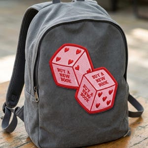 May include: A charcoal gray canvas backpack featuring two embroidered pink dice patches. Each die has red heart pips and the red embroidered text "BUY A NEW BOOK" on its visible faces. The backpack includes a top handle, adjustable straps, and a zipper.