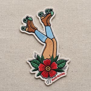 May include: An embroidered patch featuring two legs wearing light blue socks and brown roller skates with green wheels, emerging from a large red flower with green leaves. The text "SKATE BLOOMS" is stitched in red at the bottom right corner.