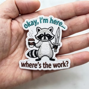 May include: An embroidered patch featuring a white raccoon with black eye markings, holding a brown coffee cup in one hand and a silver dagger in the other. The text reads "okay, i'm here..." in teal and "where's the work?" in brown thread.